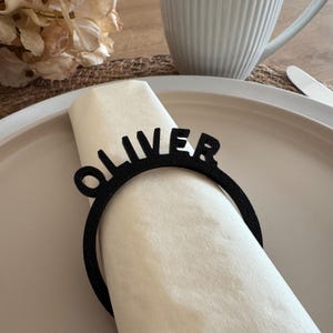 Op de afbeelding: Een zwarte servetring met de naam "Oliver" in een modern lettertype. De ring is gemaakt van duurzaam materiaal en is perfect om een persoonlijke touch aan uw tafelsetting toe te voegen.