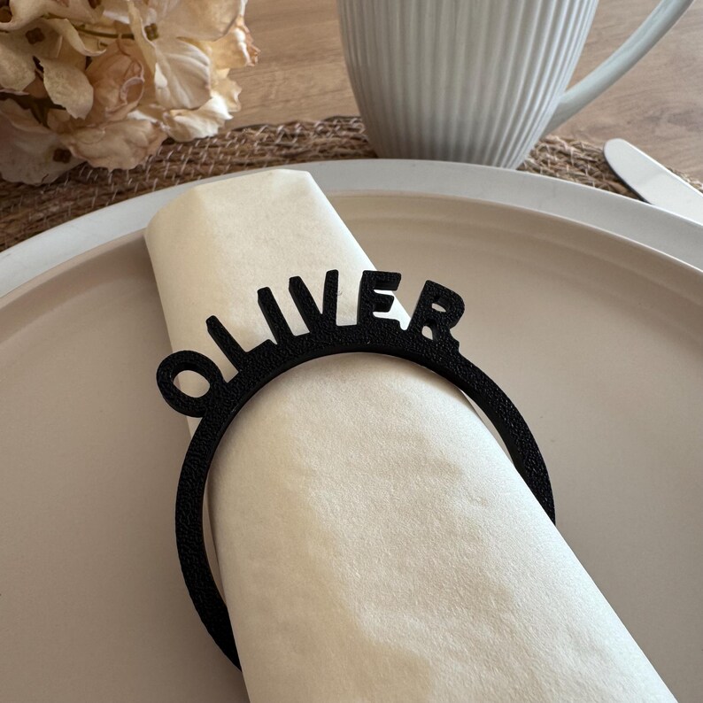 Op de afbeelding: Een zwarte servetring met de naam "Oliver" in een modern lettertype. De ring is gemaakt van duurzaam materiaal en is perfect om een persoonlijke touch aan uw tafelsetting toe te voegen.