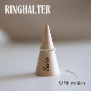 May include: A silver ring displayed on a wooden ring holder. The holder is cone-shaped with the name "Sara" engraved on it. The text "RINGHALTER" is at the top, and "NAME w&auml;hlen" is at the bottom.