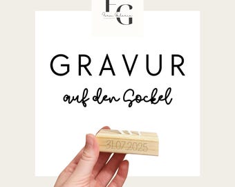 May include: A light-colored wooden block with the date "31.07.2025" engraved on it. The block is held in a hand. The text "GRAVUR auf den Sockel" is displayed in black font.