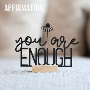 Könnte beinhalten: Ein schwarzes Holzschild mit den Worten "you are ENOUGH" in kursiver und Blockschrift, mit einem Gänseblümchen-Akzent. Das Schild ist auf einem kleinen, rechteckigen Holzsockel montiert. Das Wort "AFFIRMATION" steht über dem Schild.
