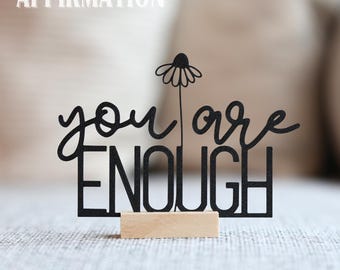 May include: A black wooden desk sign with the words "you are ENOUGH" in a cursive and block font, with a daisy accent. The sign is mounted on a small, rectangular wooden base. The word "AFFIRMATION" is above the sign.