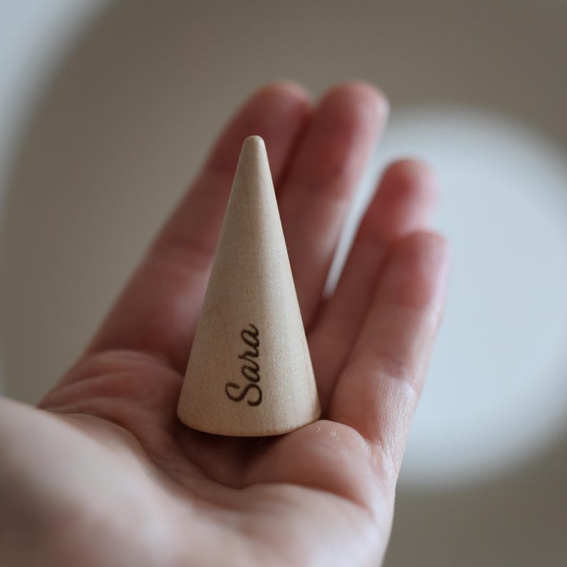 May include: A light-colored wooden ring cone with the name "Sara" engraved on the side. The cone is held in a person's hand, showcasing its smooth, tapered design. The background is softly blurred, highlighting the ring cone.