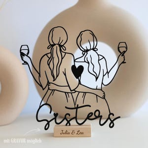Könnte beinhalten: Schwarze Metall-Silhouette von zwei Schwestern, die sich umarmen und jeweils ein Weinglas halten. Ein Herz zwischen ihnen. Das Wort "Sisters" in Schreibschrift. Ein Holzsockel mit der Aufschrift "Julia & Lea".