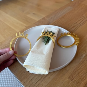 Op de afbeelding: Drie gouden servetringen met de namen "Lisa", "Mark" en "Christine" erop gedrukt. De ringen liggen op een witte servet en een lichtbruin bord.