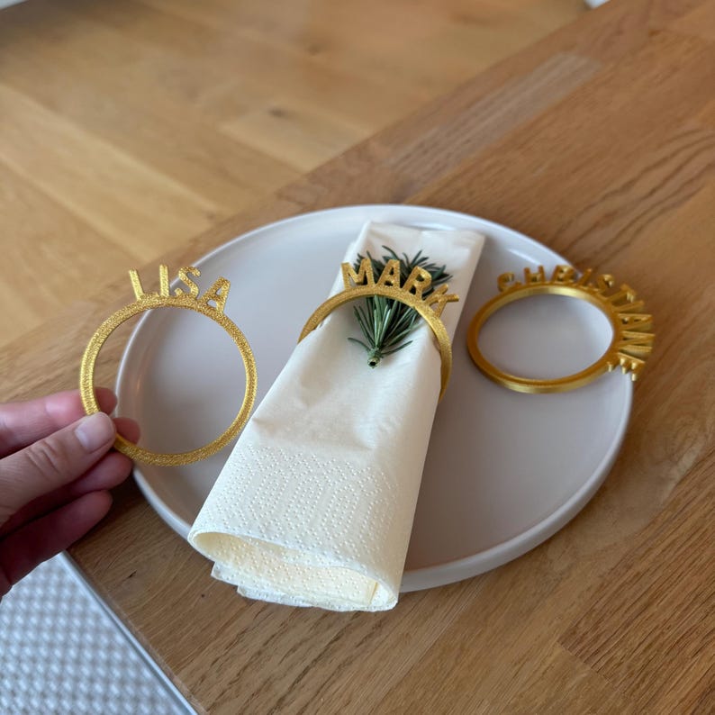 Op de afbeelding: Drie gouden servetringen met de namen "Lisa", "Mark" en "Christine" erop gedrukt. De ringen liggen op een witte servet en een lichtbruin bord.