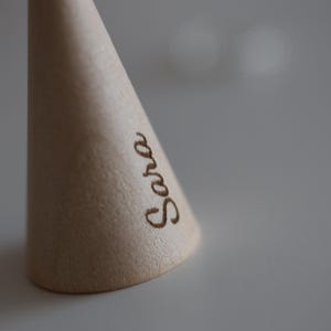 May include: A light-colored wooden cone with the name "Sara" etched in a cursive font. The cone is smooth and has a slightly rounded base. The background is a soft, neutral color.