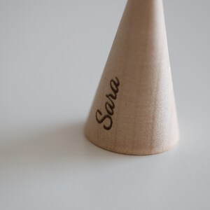 May include: A light-colored, wooden, cone-shaped object with the name "Sara" engraved on its side. The object is set against a plain, light-colored background, with the focus on the wooden cone.