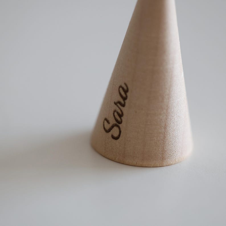 May include: A light-colored, wooden, cone-shaped object with the name "Sara" engraved on its side. The object is set against a plain, light-colored background, with the focus on the wooden cone.