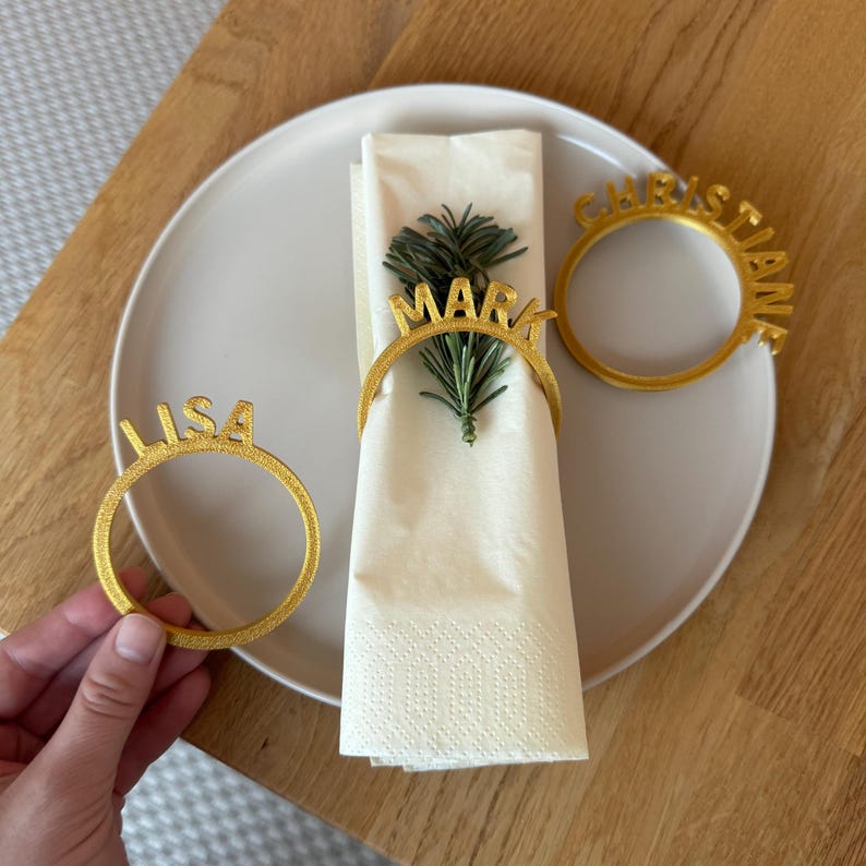 Op de afbeelding: Vier gouden servetringen met de namen "Lisa", "Mark", "Christian" en een vierde naam die gedeeltelijk verborgen is. De ringen liggen op een witte servet en een beige bord.