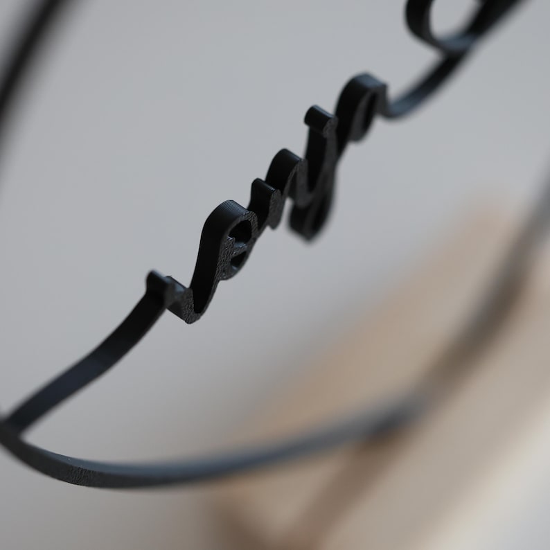 May include: A black metal decorative piece with the word "love" in cursive script. The word is integrated into a circular design, with the letters connected and forming a continuous line. The background is blurred, highlighting the word.