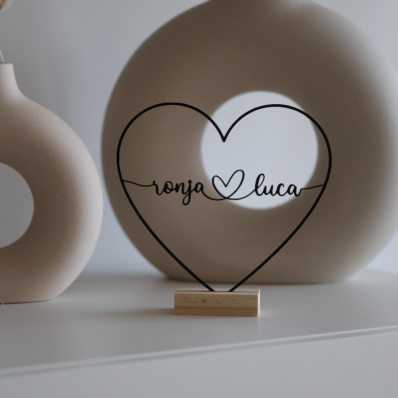 May include: A black metal heart-shaped decoration with the names "ronja" and "luca" in cursive script. The heart sits on a small wooden base. Two neutral-toned ceramic vases are in the background.