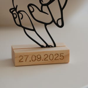 May include: A black metal art piece depicting a hand gesture, mounted on a rectangular wooden base. The base is engraved with the date 27.09.2025. The artwork is set against a neutral background.