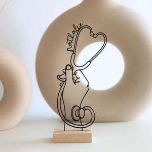 Könnte beinhalten: Eine schwarze Drahtskulptur eines Stethoskops, das von einer Hand gehalten wird, montiert auf einem kleinen Holzsockel. Das Wort "Nathalie" ist in Schreibschrift auf dem Stethoskop geschrieben. Die Skulptur steht vor einem neutralen Hintergrund mit zwei dekorativen Vasen.