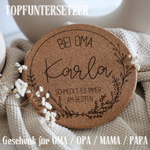 Könnte beinhalten: Ein runder, brauner Untersetzer aus Kork mit dem Namen "Karla" in Schreibschrift, umgeben von einem floralen Design. Der Text "Bei Oma" und "Schmeckt es immer am Besten" sind ebenfalls sichtbar. Die Wörter "TOPFUNTERSETZER" und "Geschenk für OMA / OPA / MAMA / PAPA" sind oben und unten.