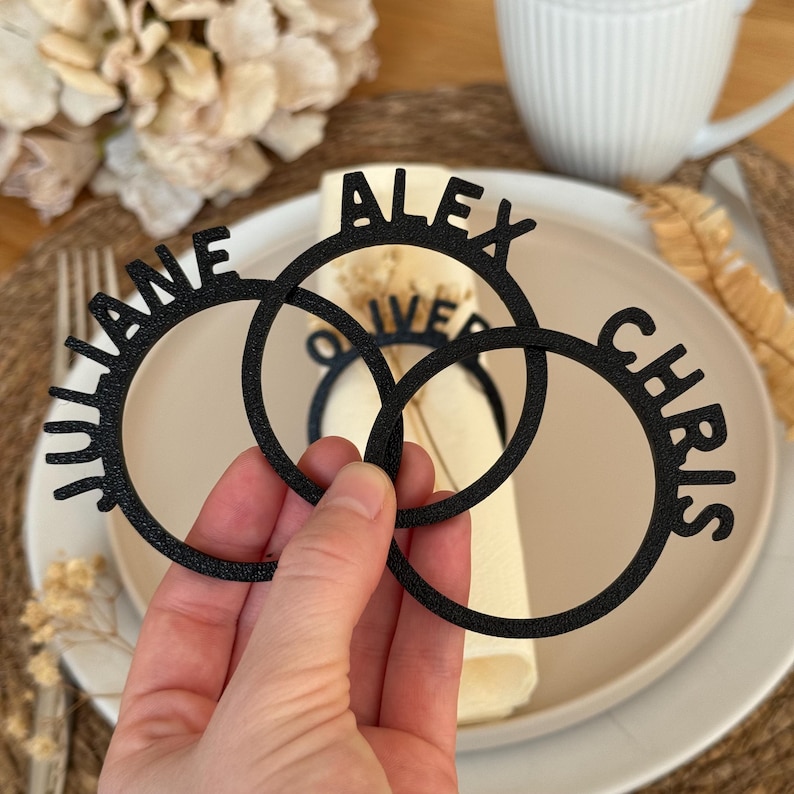 Op de afbeelding: Drie zwarte servetringen met de namen "Alex", "Chris" en "Juliane" erop. De ringen zijn in elkaar gehaakt en liggen op een witte servet.
