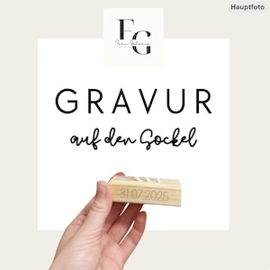 May include: A wooden block with the date "31.07.2025" engraved on it. The block is held in front of a white background with the text "GRAVUR auf den Sockel" in black. The logo "FG Farm Galerie" is in the top left corner.