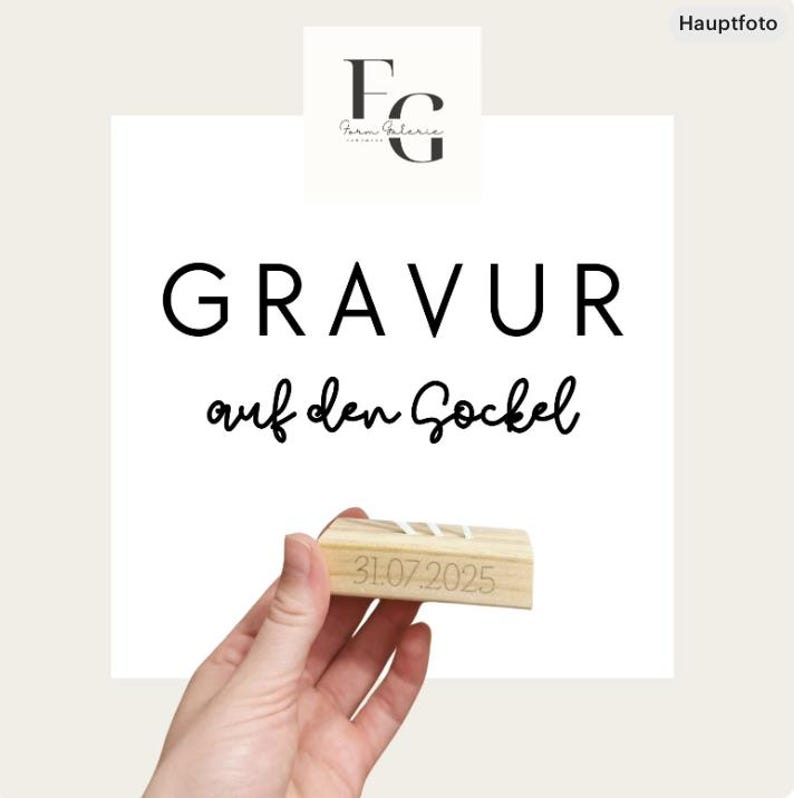 May include: A wooden block with the date "31.07.2025" engraved on it. The block is held in front of a white background with the text "GRAVUR auf den Sockel" in black. The logo "FG Farm Galerie" is in the top left corner.