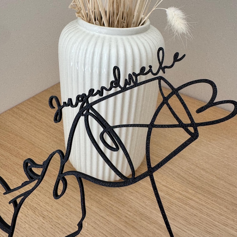 May include: A black metal decorative piece with the word "Jugendweihe" in cursive script. The design includes abstract shapes and a heart, set against a white vase with dried flowers on a wooden surface.