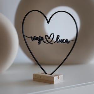 May include: A black metal heart-shaped decoration with the names "Tonja" and "Luca" intertwined, resting on a small wooden base. The heart is a wireframe design, and the names are in a cursive font. The background is neutral.