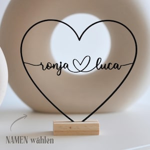 May include: A black metal heart-shaped decoration with the names "ronja" and "luca" in cursive script, connected by a heart symbol. The decoration is mounted on a small, rectangular wooden base. The text "NAMEN w&auml;hlen" is in the lower left corner.