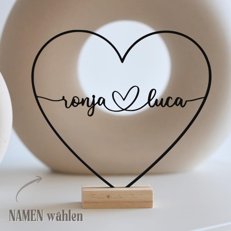May include: A black metal heart-shaped decoration with the names "ronja" and "luca" in cursive script, connected by a heart symbol. The decoration is mounted on a small, rectangular wooden base. The text "NAMEN w&auml;hlen" is in the lower left corner.