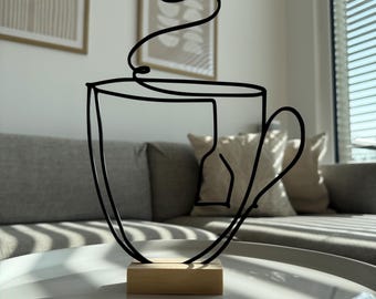 May include: A black wire sculpture of a teacup with steam rising from it. The sculpture is on a wooden base and is sitting on a white table.