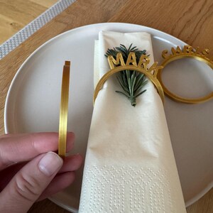 Op de afbeelding: Twee gouden servetringen met de namen "Mark" en "Carrie" erop. De ringen liggen op een witte servet met een takje groen.