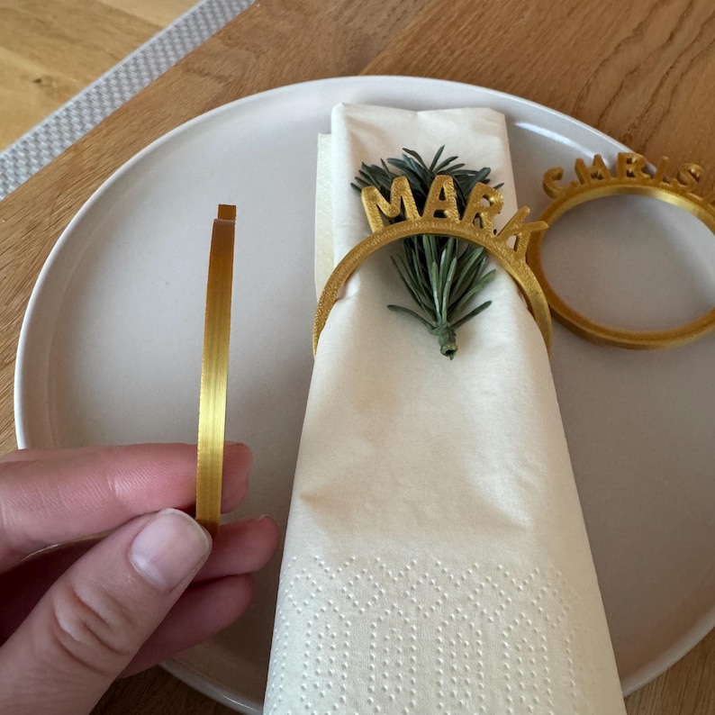 Op de afbeelding: Twee gouden servetringen met de namen "Mark" en "Carrie" erop. De ringen liggen op een witte servet met een takje groen.