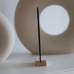 May include: A black, rectangular object is held upright by a small, natural wood block with a slot. The background features two off-white, ceramic decorative pieces. The scene is set on a white surface.