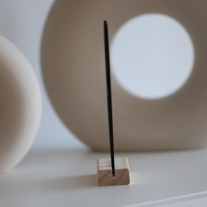May include: A black, rectangular object is held upright by a small, natural wood block with a slot. The background features two off-white, ceramic decorative pieces. The scene is set on a white surface.