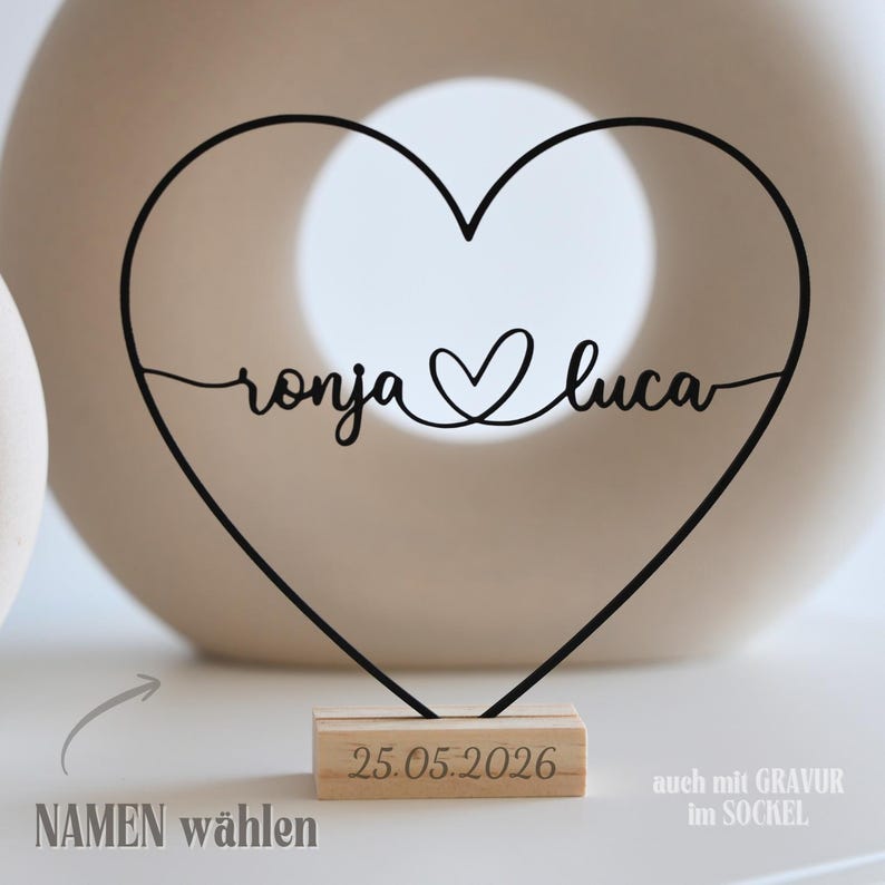 May include: A black metal heart-shaped decoration with the names "ronja" and "luca" in cursive script. The heart sits on a wooden base with the date "25.05.2026" engraved. The words "NAMEN w&auml;hlen" are also visible.