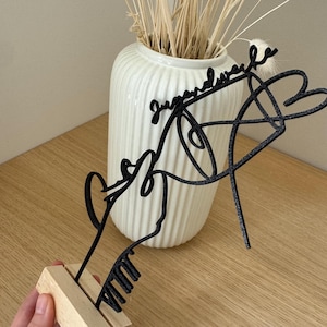 May include: A black metal sculpture with the word "Jugendwerke" in script. The sculpture depicts a stylized face, a heart, and a musical instrument, mounted on a small wooden base. A white vase with dried plants is in the background.