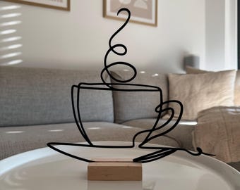 May include: A black wire sculpture of a coffee cup with steam rising from it. The sculpture is on a wooden base and is sitting on a white table.