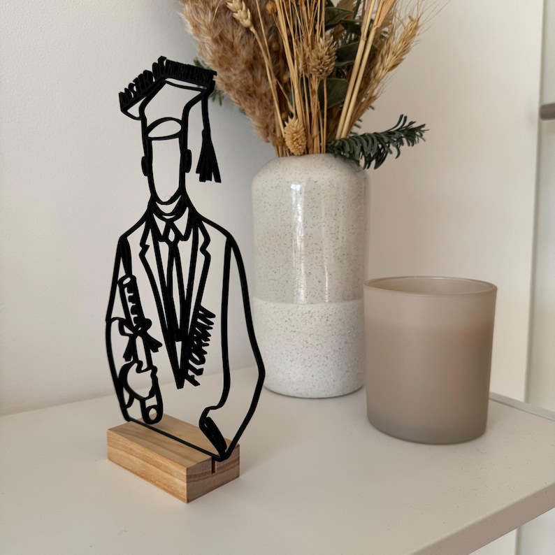 May include: Black metal silhouette of a graduate holding a diploma, standing on a wooden base. The silhouette is on a white surface with a vase and a candle holder.