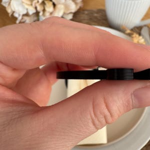 Op de afbeelding: Een zwarte plastic strik met een glad, afgerond oppervlak. De strik wordt in een hand gehouden.