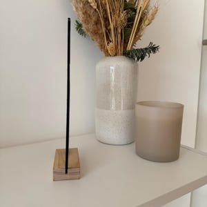 May include: A black acrylic sign with a wooden base. The sign is standing on a white shelf with a white vase and a brown candle.