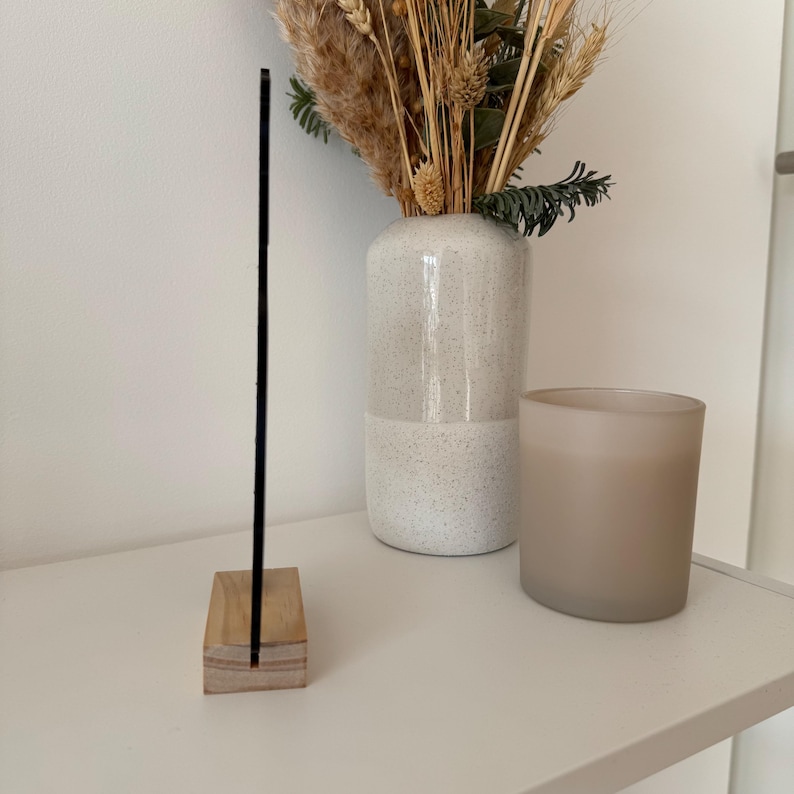 May include: A black acrylic sign with a wooden base. The sign is standing on a white shelf with a white vase and a brown candle.