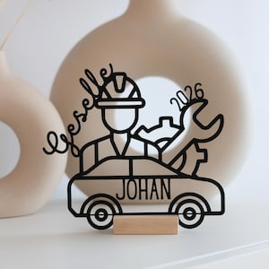 May include: Black metal car-themed decoration featuring a mechanic, wrench, and gear. The name "JOHAN" is on the car, with the year "2026" on the wrench. It's mounted on a small wooden base. A decorative item.
