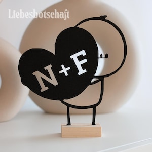 Könnte beinhalten: Eine schwarze herzförmige Dekoration mit den Buchstaben "N+F" in Weiß, gehalten von einer Strichmännchenfigur. Die Figur ist auf einem kleinen Holzsockel montiert. Das Wort "Liebesbotschaft" ist links oben aufgedruckt.