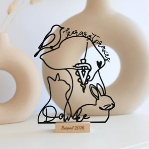 Könnte beinhalten: Eine schwarze Drahtskulptur mit den Silhouetten von Vogel, Hund, Katze und Kaninchen, einem Veterinärsymbol sowie dem Schriftzug "Tierarztpraxis" und "Danke". Die Skulptur steht auf einem rechteckigen Holzsockel mit der Aufschrift "Beispiel 2026".