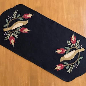 May include: A navy blue felt table runner with a scalloped edge. The runner is decorated with appliqued birds, red flowers, and green leaves. The birds are tan, and the flowers have red petals and tan centers. The leaves are green.