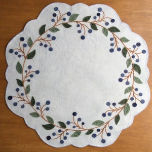 Puede incluir: Un camino de mesa de fieltro blanco con un borde festoneado presenta un dise&ntilde;o bordado de bayas azules y hojas verdes alrededor del per&iacute;metro.