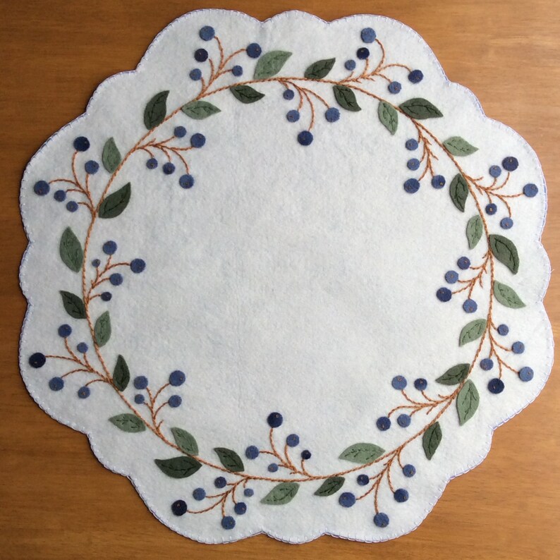 Puede incluir: Un camino de mesa de fieltro blanco con un borde festoneado presenta un dise&ntilde;o bordado de bayas azules y hojas verdes alrededor del per&iacute;metro.
