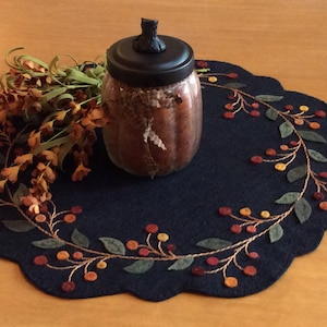 Puede incluir: Un camino de mesa de fieltro azul oscuro con un borde festoneado presenta un dise&ntilde;o bordado de una vid con hojas verdes y bayas naranjas. Un frasco de vidrio con una tapa negra se encuentra sobre el camino de mesa.
