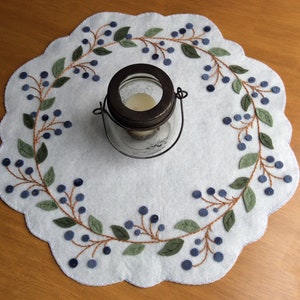 Puede incluir: Un camino de mesa de fieltro blanco con un dise&ntilde;o bordado de vid de ar&aacute;ndanos en azul y verde. Un frasco de vidrio con tapa de metal se encuentra en el centro del camino de mesa.