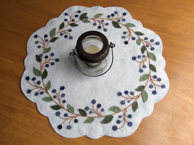 Puede incluir: Un camino de mesa de fieltro blanco con un dise&ntilde;o bordado de vid de ar&aacute;ndanos en azul y verde. Un frasco de vidrio con tapa de metal se encuentra en el centro del camino de mesa.