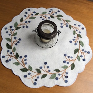 May include: White felt table runner with a blue and green embroidered design of a blueberry vine. A glass jar with a metal lid is in the center of the runner.