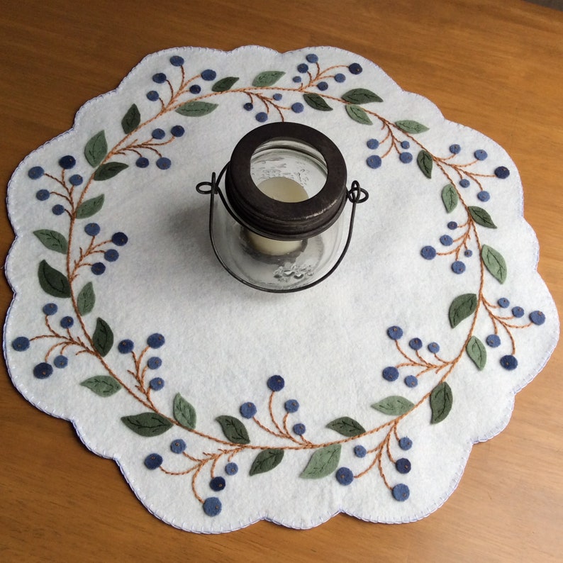Puede incluir: Un camino de mesa de fieltro blanco con un dise&ntilde;o bordado en azul y verde de una vid de ar&aacute;ndanos. Un tarro de vidrio con tapa de metal est&aacute; en el centro del camino de mesa.