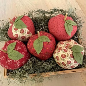 May include: A wooden basket filled with five fabric apples. The apples are primarily red with green felt leaves and brown stems. Some apples have a floral pattern. The basket is lined with dried moss.